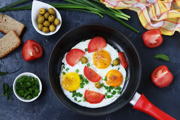 Scrambled eggs with tomatoes and green onions in a frying pan on a dark background, top view, on the table are olives, bread and tomatoes