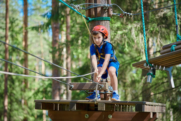 Cute boy enjoying activity in climbing adventure park at sunny summer day. Kid climbing in rope playground structure. Child in forest adventure park, extreme sport.