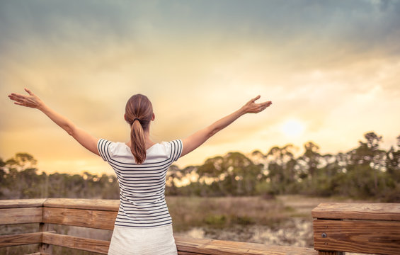 Happy young woman with arms raised facing a beautiful sunset. People nature getaway concept