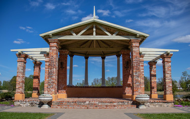 A large red brick gazebo with columns stands in a field against a blue sky and green grass
