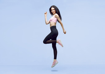 Woman in trendy sportswear jumping. Smiling beautiful slim brunette young girl in fashion leggings and pink top expressing happy emotions on blue background.