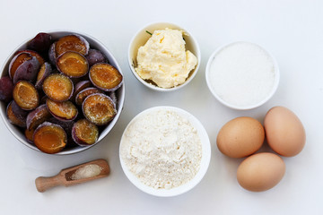 Ingredients for plum pie baking on a white background: butter, flour, sugar, baking powder, eggs, frozen plums