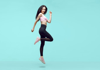 Woman in trendy sportswear jumping. Smiling beautiful slim brunette young girl in fashion leggings and pink top expressing happy emotions on blue background.