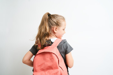 Fototapeta Back view of little girl photographed against white background wearing school uniform dress isolated holding a coral backpack on one shoulder obraz