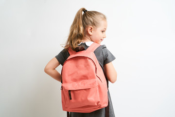 Little girl photographed against white background wearing school uniform dress isolated holding a coral backpack on both shoulders - obrazy, fototapety, plakaty