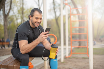 handsome man in summer outdoors on the sports ground drinking water from a bottle shaker, using a smartphone