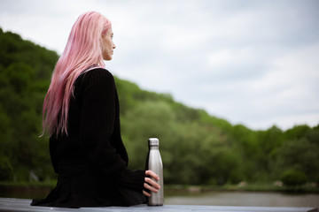 Portrait of young girl with pink hair sitting in park with thermo bottle in hand