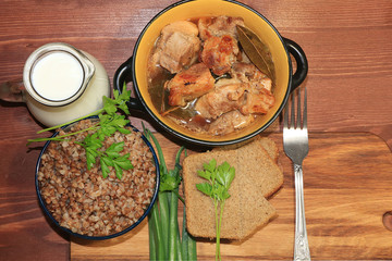 Natural organic buckwheat porridge in a clay pot, meat in a pot, a jug of milk and onions with parsley, top view. Delicious dietary breakfast of cereal and meat on wooden background.