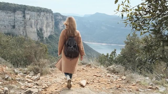 Young girl is walking among the mountains in the forest overlooking the lake, on the back is a brown hipster backpack