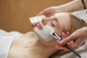 people, beauty, cosmetology and treatment concept - close up of beautiful young woman lying with closed eyes and cosmetologist applying facial mask by brush at spa