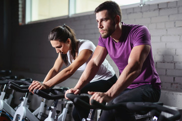 Fototapeta na wymiar Attractive woman and man biking in the gym, exercising legs doing cardio workout cycling bikes. Couple in a spinning class wearing sportswear. Fitness healthy lifestyle concept