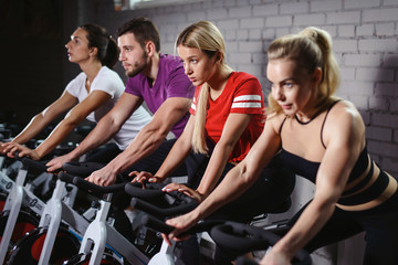 Group of smiling friends at gym exercising on stationary bike. Happy cheerful athletes training on exercise bike. Young men and woman working out at a class in the gym.