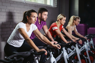 Fototapeta na wymiar Group of smiling friends at gym exercising on stationary bike. Happy cheerful athletes training on exercise bike. Young men and woman working out at a class in the gym.