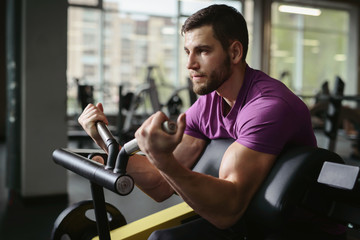 Young healthy sporty athletic man in the gym performing biceps curls with a barbell and doing arms exercises. Training in fitness studio