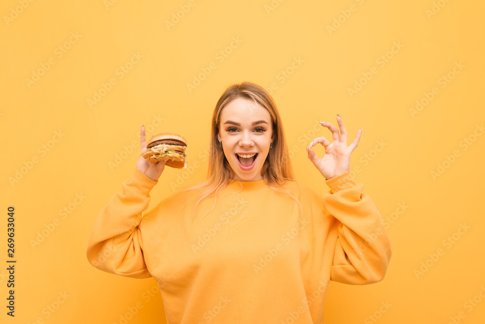 Wall mural joyful girl with a burger in her hand stands on a yellow background, shows an ok sign and looks at t