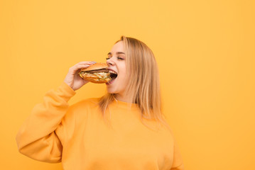 Portrait of a happy girl bites a delicious large burger with her eyes closed on a yellow...