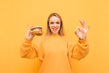 Happy girl shows an OK sign and looks in camera and holds a burger in his hand, isolated on a yellow background. Hungry girl eats fast food. Student eats fast food. Ok sign.