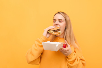Closeup portrait of a girl biting a burger with her eyes closed on a yellow background, wearing...