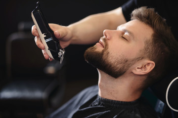 Getting perfect shape. Close-up side view of young bearded man getting beard haircut by hairdresser...