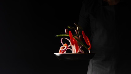 Chef preparing pepperoni sausages with green beans, sweet bell peppers and red onion rings, on a black background, a recipe book, German sausages, freeze in motion