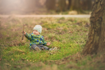 Small boy is sittin on grass and handling stick in spring park near big tree