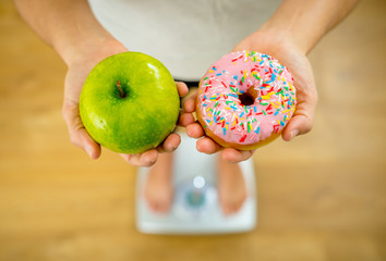 Woman on scale measuring weight holding apple and donuts choosing between healthy or unhealthy food