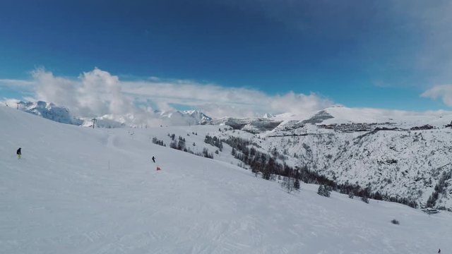 Aerial view of winter landscape at a mountain ski resort with people skiing and snowboarding down the slope. Drone flying uphill with view of clouds and mountain peaks in the distance. Winter sports.