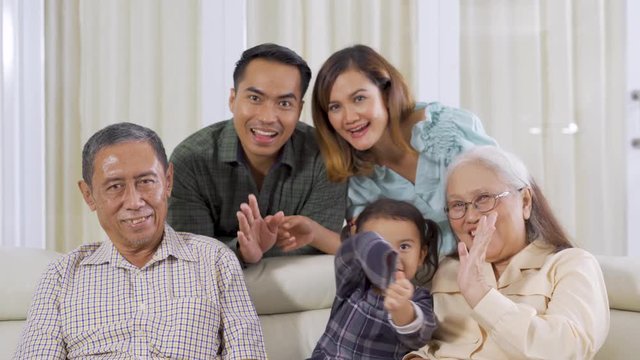 Happy three generation family smiling at the camera while sitting on the sofa and waving hands together. Shot in 4k resolution