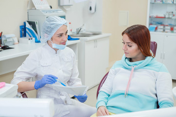 Young beautiful female dentist talking to a patient in the office. A red-haired woman sits in a chair with an orthodontist and listens to appointments. Dental services
