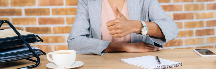 Obraz na płótnie Canvas panoramic shot of Casual african american businesswoman at desk with coffee and notebook showing thumb up