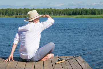 a man in a hat, sitting on the pier and looking into the distance, lying next to the fishing rod for fishing, against the blue sky and the lake