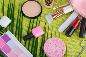 on a light green background and palm leaves, scattered in a pink cosmetic bag makeup items, brush, nail Polish, lipstick, powder, eye shadow and sponge to cleanse the face