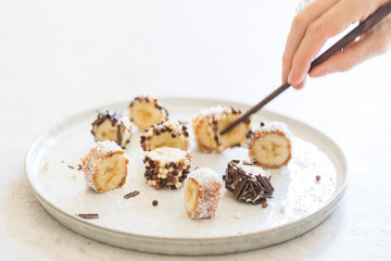Hand holding wooden chopsticks with banana maki rolled in almond, cashew and peanut butter and covered with chocolate beads, shavings and coconut powder served on a white plate. Horizontal, close up.  © Delphine