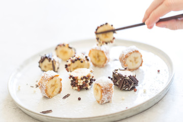 Hand holding wooden chopsticks with banana maki rolled in almond, cashew and peanut butter and covered with chocolate beads, shavings and coconut powder served on a white plate. Horizontal, close up. 