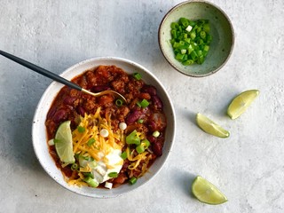 Top view of one bowl of homemade organic chili con carne served with grated cheddar cheese, scallions and sour cream, extra scallions and lime wedges. Traditional Mexican dinner. 