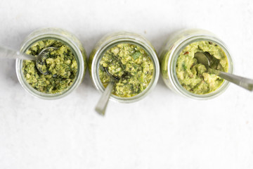 Top view of three pestos in glass jars: avocado, hemp seeds and pumpkin seeds pesto; almond, mint and basil pesto: walnut, kale and parsley pesto served with a spoon on light background. Flat lay.