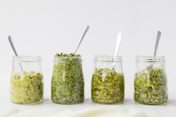 Four pestos in glass jars: avocado, hemp seeds and pumpkin seeds pesto; almond, mint and basil pesto: walnut, kale and parsley pesto served with a spoon on light background. Horizontal, copy space