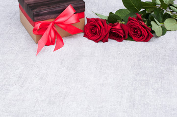 A beautiful gift box with a red ribbon along with fresh red roses and empty space for your design or montage. The concept of holiday and gifts.