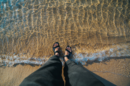 Close Up Shot Of Men's Feet At The Beach With Sea Water Ripple Effect.