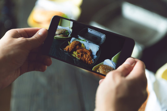 Taking photo of hamburger, french fries and fried chicken in takeaway containers with smart phone. Food delivery, fast food and food blogger concept
