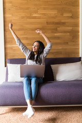 Young woman raising her hands up on the couch with laptop at home