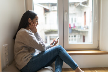 Young woman sitting listening music in wireless headphones on windowsill,Looking out the window, missing or thinking to somebody or something