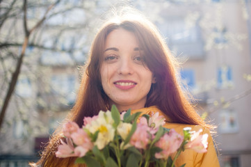 Portrait of attractive and beautiful young woman in yellow jacket holding a big bouquet of colorful flowers outdoors near the buildings on the background