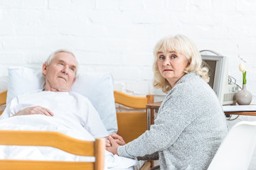 Fototapeta premium worried senior woman sitting near ill husbend and holding his hand in hospital