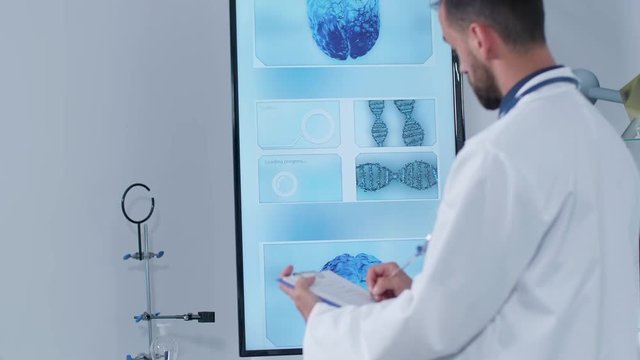 Handheld shot of doctor in his modern research center taking notes on a clipboard while a 3D brain scan is displayed on the TV in the background.