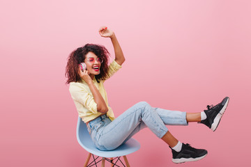 Slim african woman with long legs listening music and laughing. Stunning black female model in black shoes sitting on blue chair in pink studio.