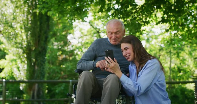 Slow motion of  happy granddaughter and grandfather in a wheelchair are having fun to look their photos or navigating in internet in cellular phone in a green park on a sunny day