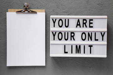 'You are your only limit' words on a modern board, clipboard with blank sheet of paper over concrete background, overhead view.