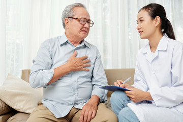 Obraz premium Senior man consulting with his doctor and she writing a prescription on the clipboard while they sitting on sofa at home