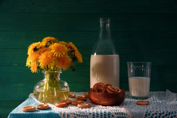 Dandelions and milk. Rustic still life. Bouquet of dandelions, the milk in the bottle and bagels. Green wooden background.